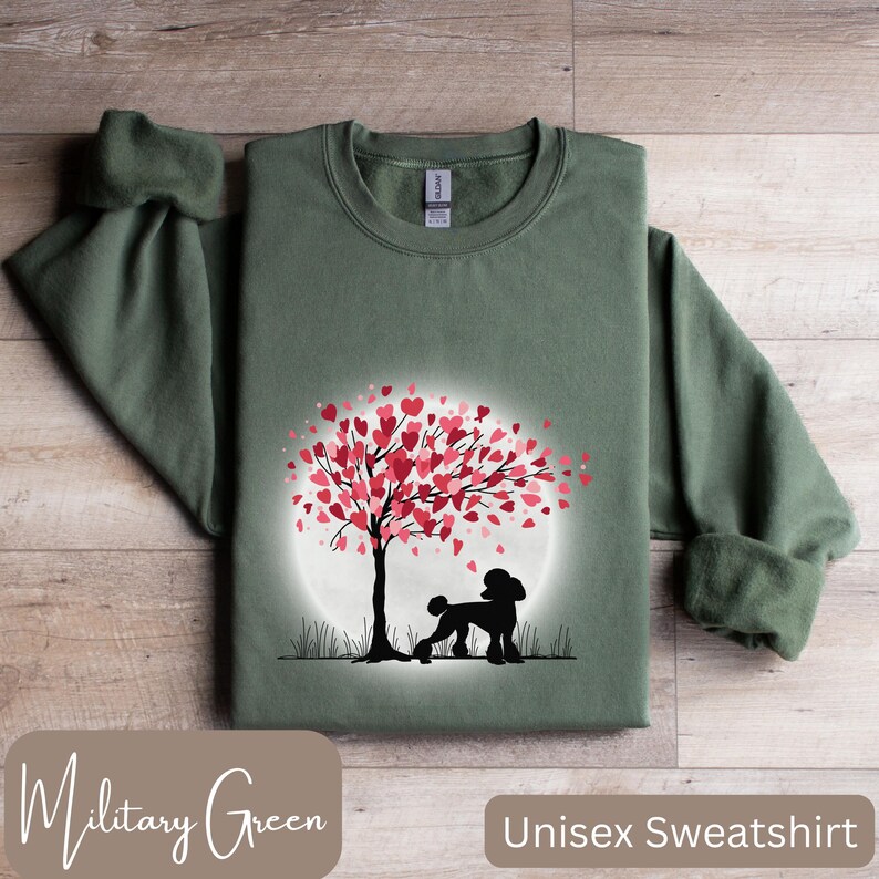 Op de afbeelding: Een unisex sweatshirt in militaire groene kleur met een silhouet van een poedel die onder een boom staat met roze harten als bladeren. De boom is omringd door een witte cirkel.