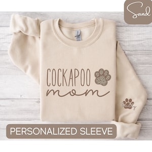 May include: A beige sweatshirt with the text "Cockapoo mom" in brown lettering. A paw print with a leopard print design is next to the text. The sleeve has a smaller paw print with the text "Buddy" in brown lettering.