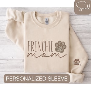 May include: Beige sweatshirt with the text "Frenchie mom" and a paw print with a leopard print design. The sleeve has a smaller paw print with the text "Buddy".