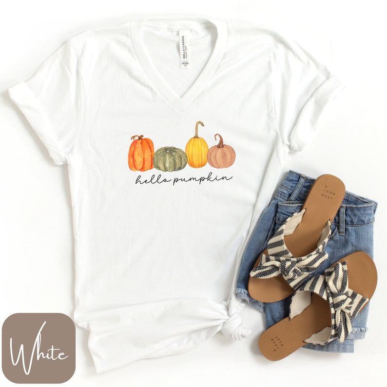 May include: White V-neck t-shirt with a watercolor design of four pumpkins in shades of orange, green, yellow, and brown. The text "hello pumpkin" is written in a script font below the pumpkins. The shirt is paired with blue jeans and striped sandals.