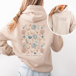 K&ouml;nnte beinhalten: Ein beiger Hoodie mit einem Muschel- und Korallendesign auf der R&uuml;ckseite. Der Text "The ocean is calling and I must go" ist auf der R&uuml;ckseite des Hoodies aufgedruckt.