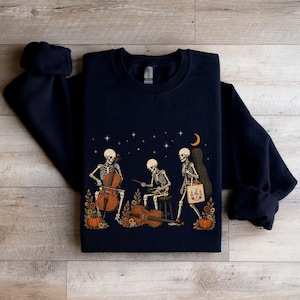May include: A dark navy blue crewneck sweatshirt featuring a graphic print of three skeletons playing string instruments, including a cello and a violin, surrounded by orange pumpkins, autumn flowers, and a crescent moon against a starry night sky.