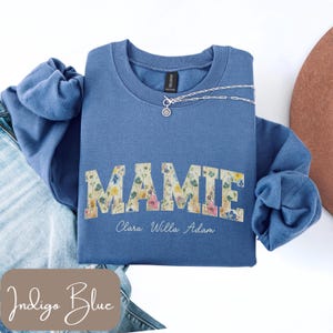 May include: Indigo blue sweatshirt featuring the word "MAMIE" in a floral design, with the names "Clara Willa Adam" underneath. A silver necklace with a pendant is on the sweatshirt.