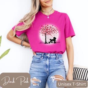 Op de afbeelding: Een roze unisex t-shirt met een afbeelding van een poedel die onder een boom staat met roze hartvormige bladeren. De boom staat voor een volle maan.