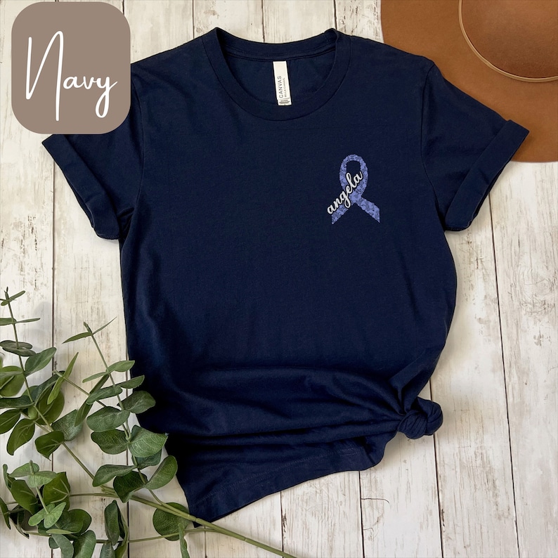 May include: Navy blue t-shirt with a purple ribbon and the word "angela" written on it.