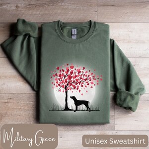 May include: A unisex sweatshirt in military green featuring a silhouette of a dog standing under a tree with pink hearts for leaves. The tree is in front of a full moon.