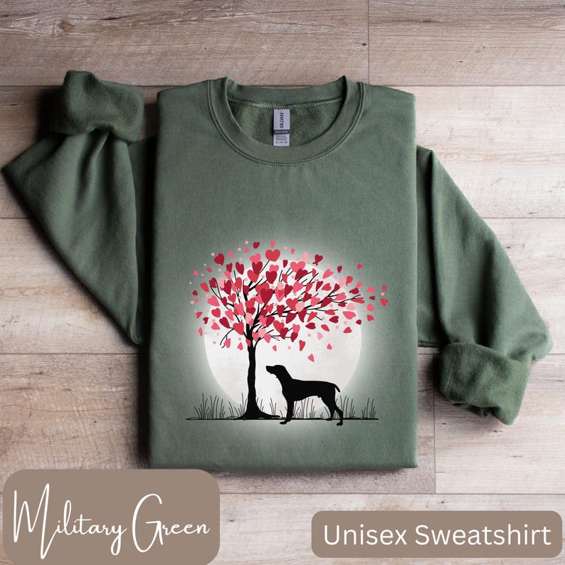 May include: A unisex sweatshirt in military green featuring a silhouette of a dog standing under a tree with pink hearts for leaves. The tree is in front of a full moon.