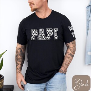 May include: Black t-shirt with the word "PAPI" in white, soccer ball-patterned letters. The sleeve has text and numbers. The shirt is worn by a person with tattoos on their arms.