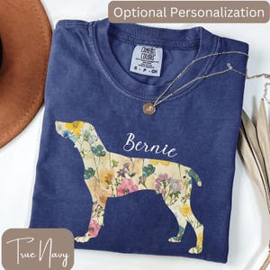 May include: A navy blue t-shirt with a floral dog silhouette and the name "Bernie" in white script. The shirt has a crew neck and short sleeves. A gold necklace with a pendant is draped across the shirt.