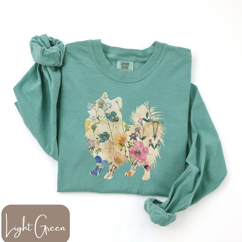 May include: Light green long-sleeve shirt with a floral dog design. The dog silhouette is filled with colorful flowers in shades of pink, yellow, blue, and white. The shirt is made of a soft material and has the Comfort Colors brand label.