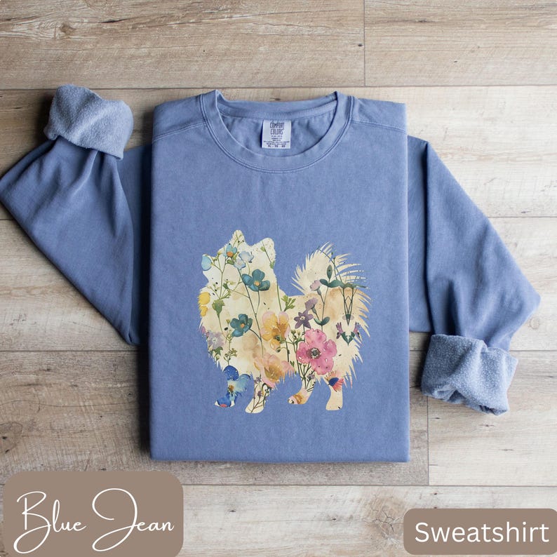 May include: A blue jean colored sweatshirt featuring a floral dog design. The dog silhouette is filled with colorful flowers. The sleeves are cuffed. The word "Sweatshirt" is in a brown box in the lower right corner.