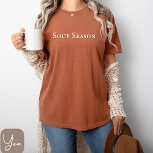 Puede incluir: Una camiseta color &oacute;xido con el texto blanco "SOUP SEASON". La camiseta se combina con vaqueros azules y una taza blanca. La est&eacute;tica general sugiere un estilo acogedor con tem&aacute;tica oto&ntilde;al.