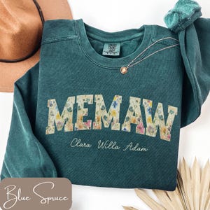 May include: A teal-colored sweatshirt with the word "MEMAW" in a floral design across the front. Below the word are the names "Clara Willa Adam" in a script font. A brown hat and a necklace are visible.