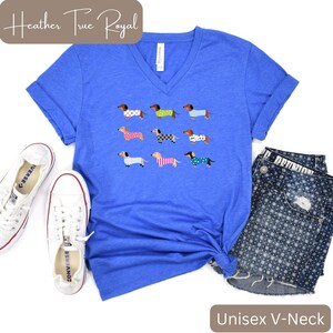 May include: A blue unisex V-neck t-shirt featuring a graphic of nine dachshunds in patterned sweaters. The text "Heather True Royal" is at the top left, and "Unisex V-Neck" is at the bottom right. White trainers and denim shorts are also in the image.