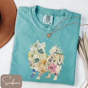 May include: Seafoam green Comfort Colors t-shirt featuring a floral dog design. The dog is filled with colorful flowers. A gold necklace with a pendant is on the shirt. The shirt is size small.