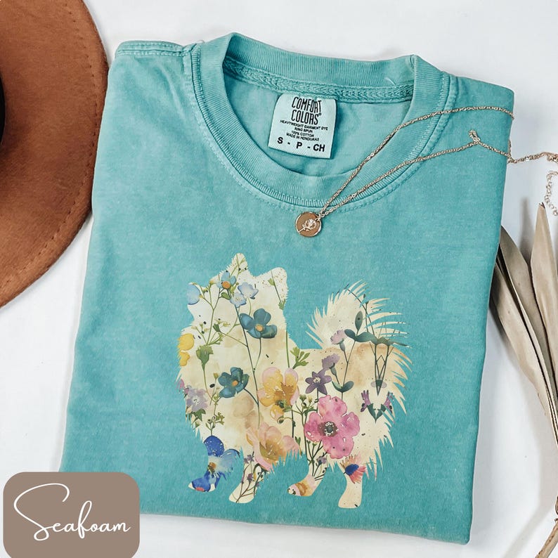 May include: Seafoam green Comfort Colors t-shirt featuring a floral dog design. The dog is filled with colorful flowers. A gold necklace with a pendant is on the shirt. The shirt is size small.