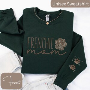 May include: A dark green unisex sweatshirt with the text "FRENCHIE mom" in brown lettering. A brown paw print with a leopard print design is next to the text. The right sleeve has the names "Cooper" and "Penny" in brown lettering with a paw print next to each name.