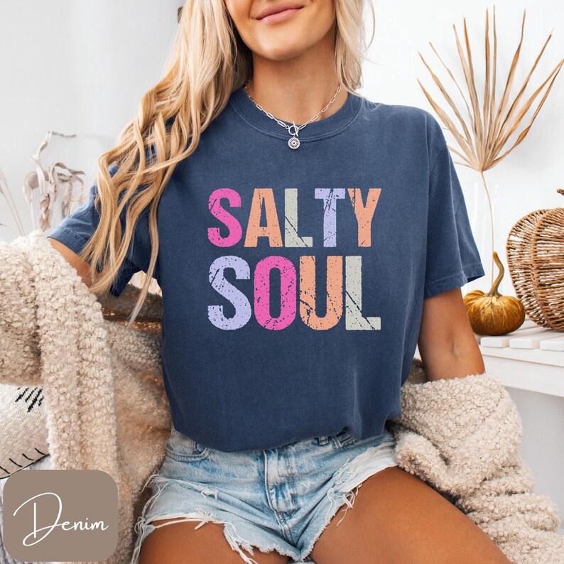 May include: A navy blue t-shirt with the words "SALTY SOUL" in a distressed, multi-colored font. The text is stacked, with each word in a different color: pink, purple, peach, and white. The shirt is paired with denim shorts.