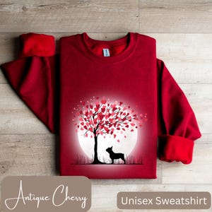May include: Red unisex sweatshirt with a graphic of a silhouette of a dog standing under a tree with pink and red hearts for leaves. The tree is in front of a white moon. The text "Antique Cherry" is at the bottom left and "Unisex Sweatshirt" is at the bottom right.