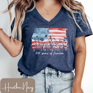 以下が含まれることがあります： ヘザーネイビーのVネックTシャツ。水彩画風のアメリカ国旗のデザインで、赤と白のストライプ、青色の背景、花のアクセントが特徴です。「250 years of freedom」の文字が旗の下にプリントされています。