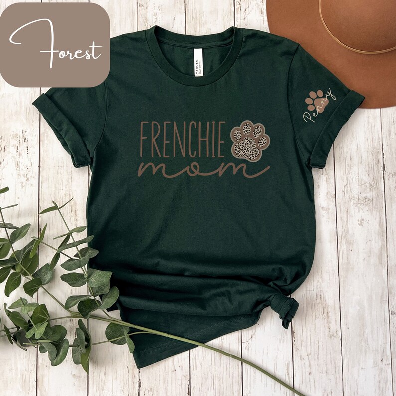 May include: A dark green t-shirt with the text "FRENCHIE mom" and a brown paw print with a leopard print design. The text "Penny" is on the sleeve of the shirt.