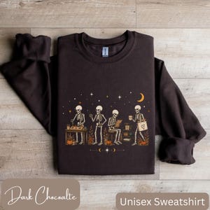May include: A dark chocolate brown crewneck sweatshirt featuring a graphic print of four skeletons engaged in various activities, including working at a desk and holding a tote bag labeled OT, surrounded by stars, a crescent moon, and pumpkins.
