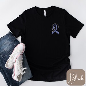 May include: Black t-shirt with a purple ribbon and the text "I wear purple for my dad". The shirt is displayed on a white background with a pair of white sneakers and blue jeans.