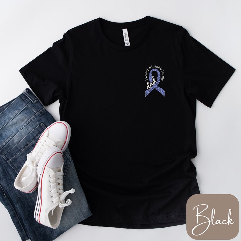 May include: Black t-shirt with a purple ribbon and the text "I wear purple for my dad". The shirt is displayed on a white background with a pair of white sneakers and blue jeans.
