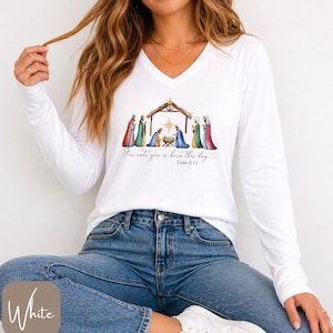 May include: A white long-sleeve V-neck shirt featuring a colorful nativity scene graphic with a stable, figures, and a star. Below the illustration, the text reads, "For unto you is born this day... Luke 2:11" in a black cursive and print font.