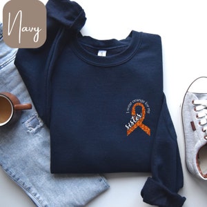 Puede incluir: Sudadera azul marino con el texto "I wear orange for my sister" y un diseño de cinta naranja. La sudadera está doblada sobre un par de vaqueros azules. También se ven una taza marrón y zapatillas grises.