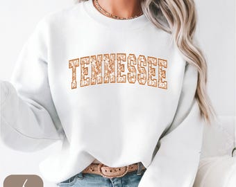 Könnte beinhalten: Weißes Sweatshirt mit dem Wort "TENNESSEE" in einem floralen Design auf der Brust. Der Text ist in einem warmen Orangebraun gehalten. Das Sweatshirt hat lange Ärmel und einen Rundhalsausschnitt.