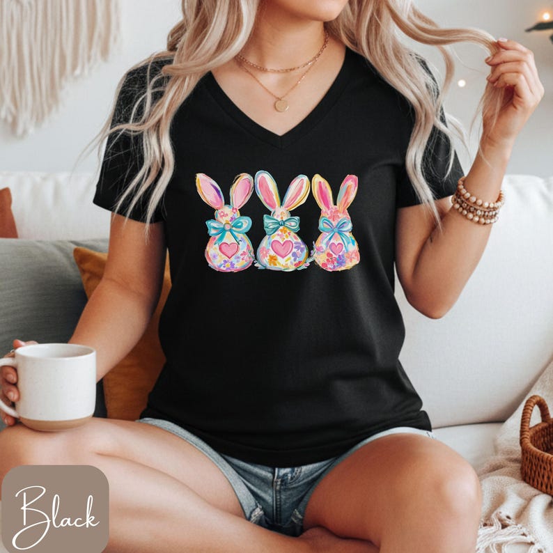 Op de afbeelding: Zwart V-hals T-shirt met drie aquarel konijn ontwerpen. Elk konijn heeft een bloemenlichaam, een hart en een strik. De konijnen zijn in tinten roze, blauw, geel en paars.