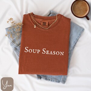 Puede incluir: Camiseta color &oacute;xido con cuello redondo y el texto "Soup Season" en blanco. La camiseta est&aacute; colocada sobre unos vaqueros azul claro. Tambi&eacute;n se ven un collar dorado y una taza de caf&eacute;.