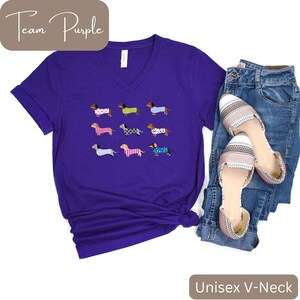 May include: A purple unisex V-neck t-shirt with a pattern of dachshunds in sweaters. The text "Team Purple" is in a brown box. The shirt is styled with blue jeans and patterned flats. The text "Unisex V-Neck" is in a brown box.
