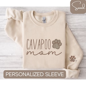 Könnte beinhalten: Ein beiger Pullover mit dem Text "CAVAPOO mom" in braunen Buchstaben und einem Pfotenabdruck mit Leopardenmuster. Der Ärmel hat einen personalisierten Pfotenabdruck mit dem Text "Buddy" in braunen Buchstaben.