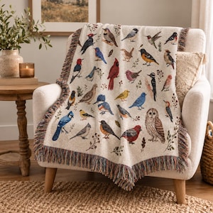 May include: A cream-coloured woven throw blanket featuring a repeating pattern of various North American bird species in shades of red, blue, yellow, brown, and black. The blanket has a textured surface and decorative fringed edges on all sides.