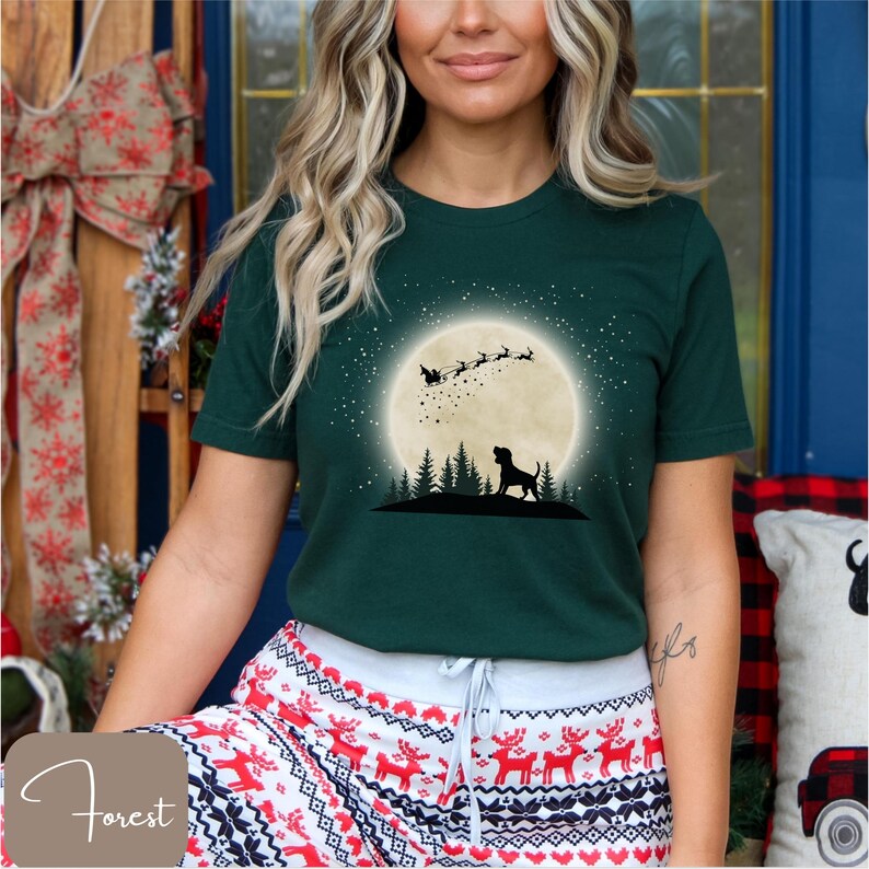 May include: Forest green t-shirt featuring a silhouette of a dog in front of a full moon with Santa and reindeer. The shirt has a graphic design of a dog, trees, and stars. The model is wearing Christmas-themed pajama pants.