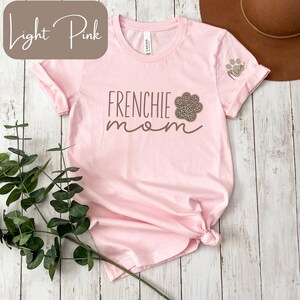 May include: Light pink t-shirt with the text "Frenchie mom" and a paw print with a leopard print design. The shirt has a small embroidered paw print with the name "Mia" on the sleeve.