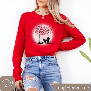 Op de afbeelding: Rood longsleeve t-shirt met een afbeelding van een poedel die onder een boom staat met roze harten als bladeren. De boom staat voor een volle maan.