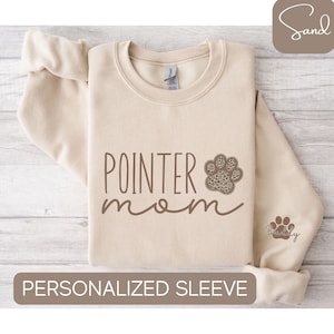 May include: A beige sweatshirt with the text "POINTER mom" in brown lettering. A brown paw print with a leopard print design is next to the text. The text "Buddy" is on the sleeve of the sweatshirt.