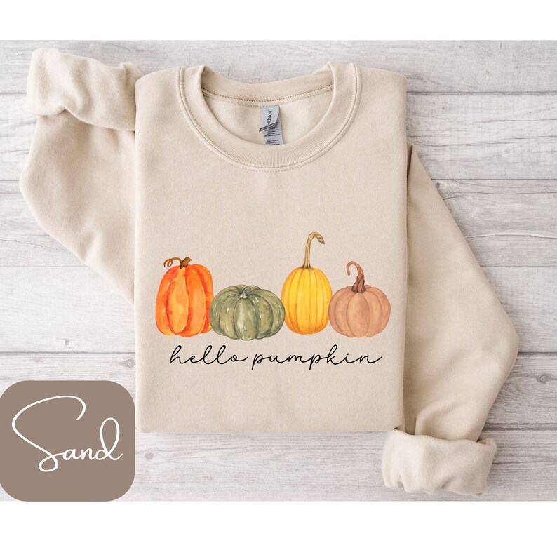 May include: Sand-colored sweatshirt featuring a watercolor design of four pumpkins in orange, green, and yellow. The phrase "hello pumpkin" is written in a black script font below the pumpkins. The sweatshirt has a crew neck and long sleeves.