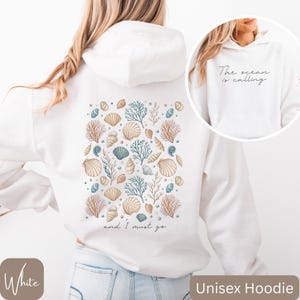K&ouml;nnte beinhalten: Wei&szlig;er Unisex-Hoodie mit einem Muschel- und Korallendesign auf der R&uuml;ckseite. Der Text "and I must go" ist unter dem Design gedruckt. Die Vorderseite des Hoodies hat den Text "The ocean is calling" auf der Vorderseite.