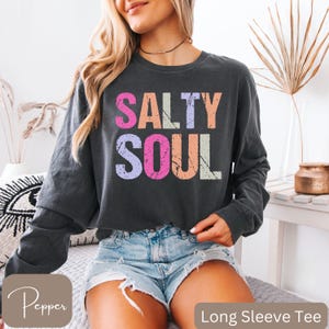 May include: Dark gray long sleeve tee with the words "SALTY SOUL" in colorful, distressed lettering. The shirt is paired with denim shorts. The words "Long Sleeve Tee" are at the bottom of the image.