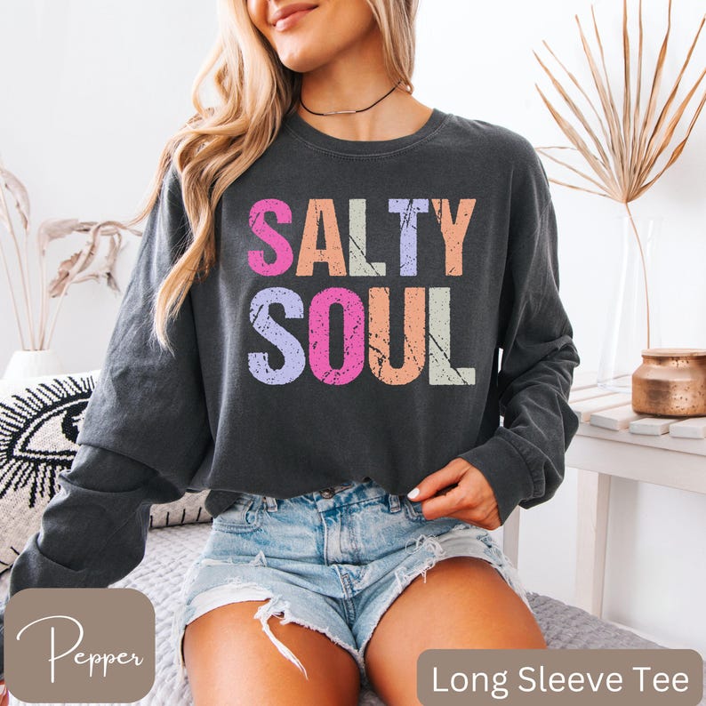 May include: Dark gray long sleeve tee with the words "SALTY SOUL" in colorful, distressed lettering. The shirt is paired with denim shorts. The words "Long Sleeve Tee" are at the bottom of the image.