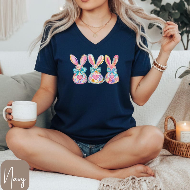 Op de afbeelding: Marineblauwe V-hals T-shirt met een aquarelontwerp van drie paashazen met kleurrijke strikken en bloemmotieven. De konijnen staan in het midden van het shirt. Het T-shirt is gemaakt van zacht materiaal.