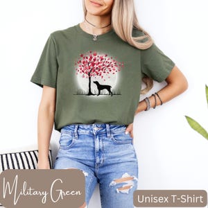 May include: A unisex t-shirt in military green featuring a silhouette of a dog standing under a tree with pink heart-shaped leaves. The tree is in front of a full moon. The text "Military Green" is at the bottom left of the image and "Unisex T-Shirt" is at the bottom right.