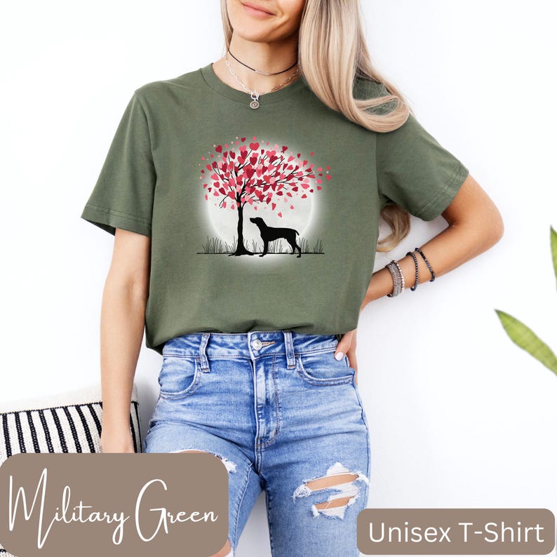 May include: A unisex t-shirt in military green featuring a silhouette of a dog standing under a tree with pink heart-shaped leaves. The tree is in front of a full moon. The text "Military Green" is at the bottom left of the image and "Unisex T-Shirt" is at the bottom right.