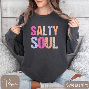 May include: Dark gray sweatshirt with the words "SALTY SOUL" in colorful, distressed lettering. The text is in pink, orange, purple, and white. The sweatshirt is a crew neck style.