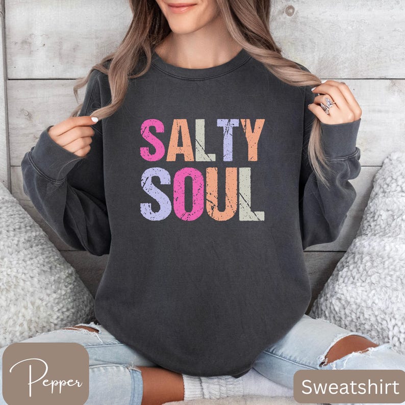 May include: Dark gray sweatshirt with the words "SALTY SOUL" in colorful, distressed lettering. The text is in pink, orange, purple, and white. The sweatshirt is a crew neck style.