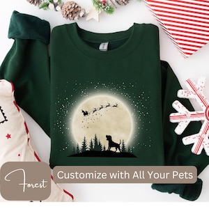 May include: A dark green sweatshirt featuring a silhouette of a dog in front of a full moon with Santa's sleigh and reindeer. The text "Customize with All Your Pets" is visible. The sweatshirt is surrounded by holiday decorations.